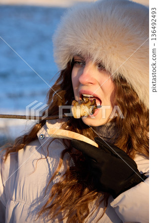 Young woman in a fur hat enjoys grilled bacon and bread outdoors, in winter. 136492493