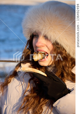 A girl in a fur hat eats lard and bread on a skewer in winter, with snow visible in the 136492494