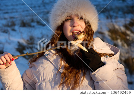 Young woman eating a grilled marshmallow on a stick outside in winter with a snowy scene. Young woman eating a grilled marshmallow on a stick outside in winter with a snowy scene. 136492499