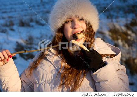 Girl in Fur Hat and Winter Coat Enjoying Grilled Snack on a Stick Outdoors in Snowy Weather. Girl in Fur Hat and Winter Coat Enjoying Grilled Snack on a Stick Outdoors in Snowy Weather. 136492500