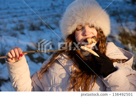 Young woman in winter hat enjoys grilled vegetables on a stick in the snowy nature. Young woman in winter hat enjoys grilled vegetables on a stick in the snowy nature. 136492501