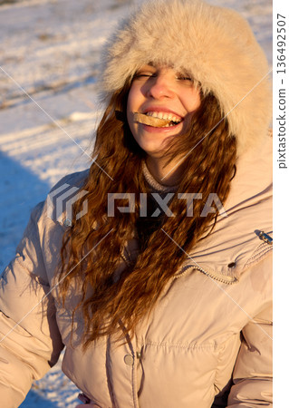 Bright smile of a young woman with a fur hat, enjoying bread in winter sun. 136492507