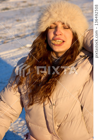Young woman with closed eyes, enjoying snack in the winter, wears a fluffy hat and a warm jacket. 136492508
