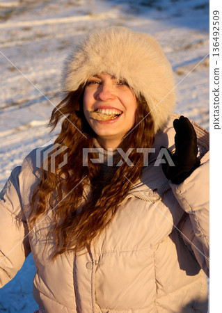 A cheerful young woman smiles while savoring bread in a winter scene, wearing a fur hat. 136492509
