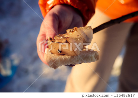 Close-up of a hand in orange jacket with marshmallow on a stick over bread, lit by sunlight. 136492617