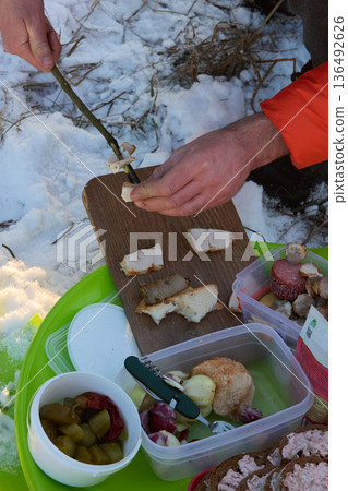 Winter picnic with a person cooking bacon on a stick outdoors with snow and food. 136492626