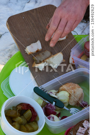 Picnic outdoors: A hand places bacon on bread, amidst food containers and a snowy landscape. 136492629