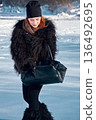 Elegant woman in fur coat and hat on snow, looking down at her black purse. 136492695