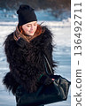 Red-haired woman with black fur coat opening a black bag in snowy winter landscape. 136492711
