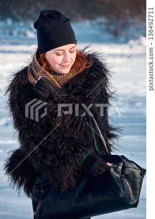 Red-haired woman with black fur coat opening a black bag in snowy winter landscape. 136492711