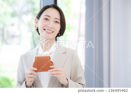 一位身穿西裝的職業女性面帶微笑，手拿錢包，在綠色背景下看向鏡頭。 136495462
