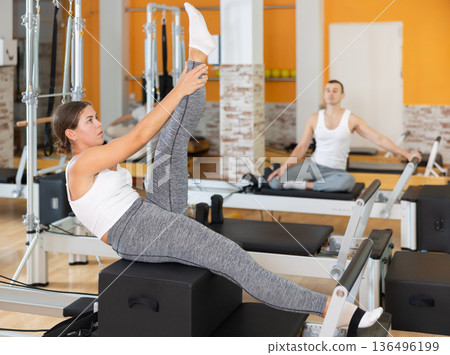 Girl works out in gym and performs set of exercises to stretch leg muscles with help of reformer Girl works out in gym and performs set of exercises to stretch leg muscles with help of reformer 136496199