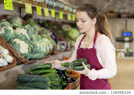 Woman seller working in supermarket and lays out cucumber on counter 136496326