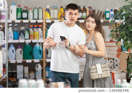 couple stands near the shelves with household chemicals in a supermarket with a phone in their hands 136496346