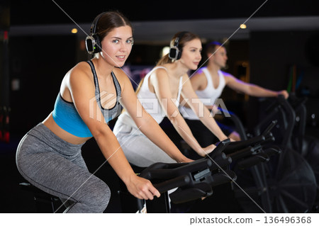 Girl in earphone performs exercises with stationary bicycle in gym 136496368