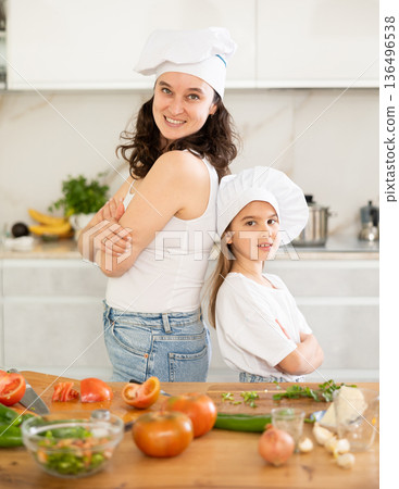 Mother and girl in white cooking hats are standing in kitchen near table Mother and girl in white cooking hats are standing in kitchen near table 136496538