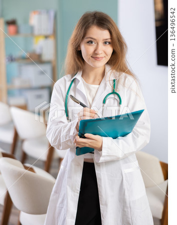 Portrait of young coach female doctor standing with folder with documents and materials in hands and talking in corporate seminar and inviting in auditorium 136496542