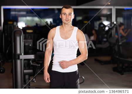 Young man trainer in sportswear poses in middle of fitness center gym. Young man trainer in sportswear poses in middle of fitness center gym. 136496571