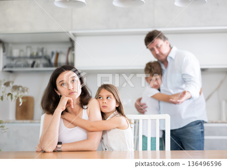 Offended mother sitting at the kitchen table being hugged by little daughter 136496596
