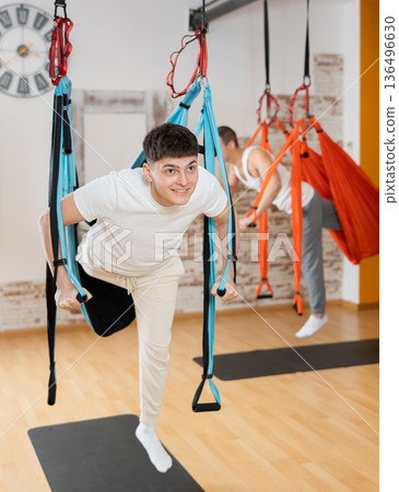 Guy practicing aerial pilates during group class in fitness center Guy practicing aerial pilates during group class in fitness center 136496630