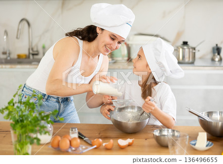Mom sifts and adds flour to bowl, girl mixes ingredients with whisk. 136496655
