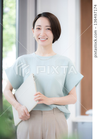 A middle-aged woman with a smile looking at the camera in an office with a beautiful green background. Spring/summer office casual wear 136497174