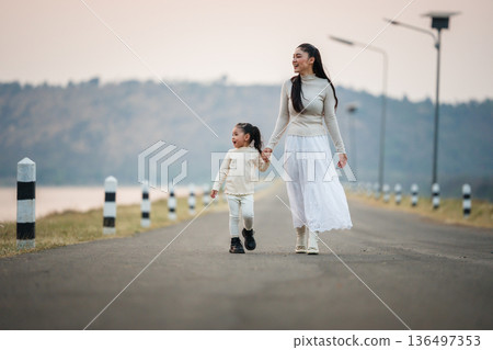 Mother and child girl holding hands walking on road by the lake at sunset. 136497353