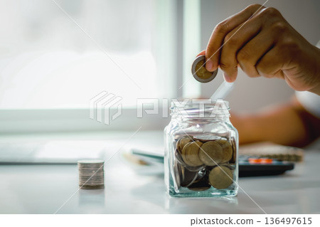 Hand putting coin into glass jar with stacks of coins and calculator, concept of money saving, financial planning, investment for future, and retirement fund. Hand putting coin into glass jar with stacks of coins and calculator, concept of money saving, financial planning, investment for future, and retirement fund. 136497615