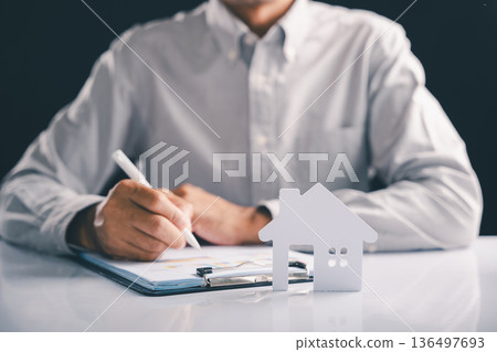 Analyzing real estate charts, person uses pen near a model house, suggesting detailed property analysis for an investment, reflecting focus on housing market and financial implications of real estate Analyzing real estate charts, person uses pen near a model house, suggesting detailed property analysis for an investment, reflecting focus on housing market and financial implications of real estate 136497693