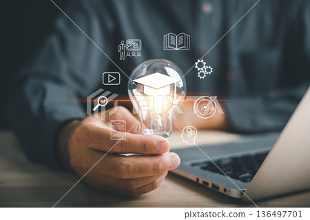 E learning, innovation, skill, idea, creativity, knowledge concepts show as a hand holds bulb with graduation cap for postgraduate studies, path to learning and education icons near laptop symbolize 136497701