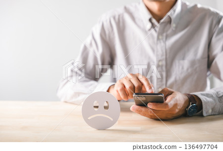Review reflecting negative customer feedback, man submits bad experience survey with smartphone, unhappy satisfaction rating shown by sad face symbol on wooden table against white background Review reflecting negative customer feedback, man submits bad experience survey with smartphone, unhappy satisfaction rating shown by sad face symbol on wooden table against white background 136497704