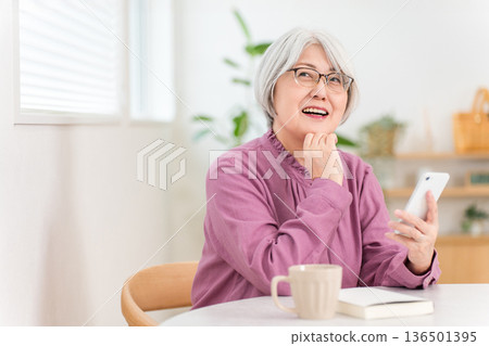 A gray-haired senior woman looking at her smartphone in the living room (search, think, imagine, excitement, idea) 136501395