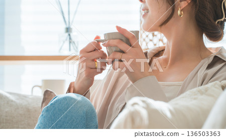 A stylish woman sitting on a sofa in the living room and drinking a hot drink A stylish woman sitting on a sofa in the living room and drinking a hot drink 136503363