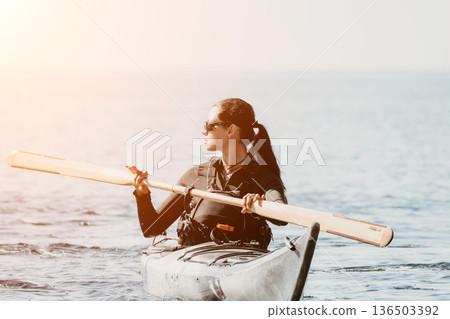 Kayaking Woman Paddle Ocean - A woman in a kayak on the ocean holding a paddle with the sun setting in the background. 136503392