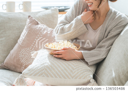 Smiling Asian woman sitting on the sofa in the living room and eating popcorn Smiling Asian woman sitting on the sofa in the living room and eating popcorn 136503486