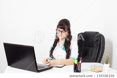 Female doctor or medical professional writing patient notes at office desk. Laptop computer, anatomical chart on wall behind. Medical work environment. Female doctor or medical professional writing patient notes at office desk. Laptop computer, anatomical chart on wall behind. Medical work environment. 136506109