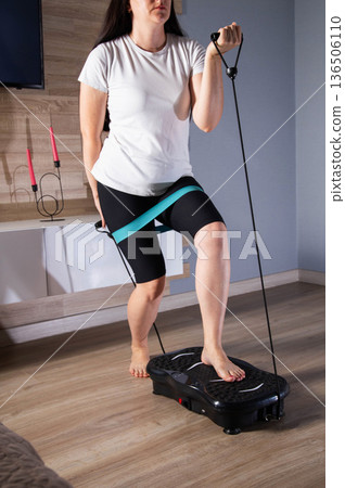 Brunette girl exercises with expanders at home on a vibration platform to reduce weight and cellulite. Exercise at home, lifestyle 136506110