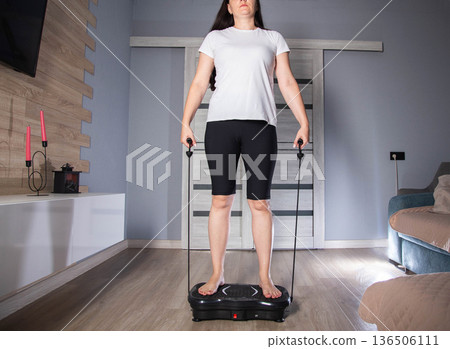 Brunette girl doing sports on vibration platform with expanders against the background of interior in the room. Weight loss and cellulite concept. Copy space for text Brunette girl doing sports on vibration platform with expanders against the background of interior in the room. Weight loss and cellulite concept. Copy space for text 136506111