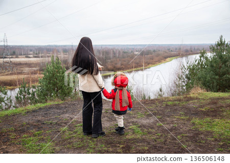 Back view of adult woman and young child holding hands, standing on hill overlooking river and vast autumn forest landscape under overcast sky, pointing finger. Back view of adult woman and young child holding hands, standing on hill overlooking river and vast autumn forest landscape under overcast sky, pointing finger. 136506148