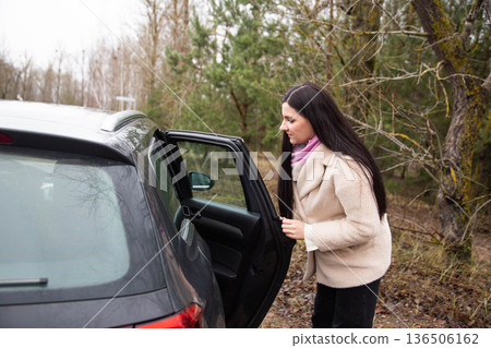 Young adult woman opening car door next to forest edge on overcast day, preparing to enter vehicle, travel and outdoor adventure concept 136506162