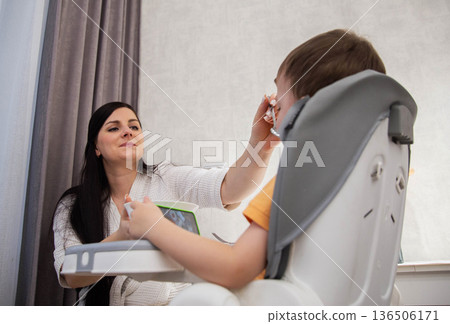 Mother feeds child sitting in high chair, child watches tablet, everyday activity of feeding time, focus on family care, comfortable indoor scene 136506171