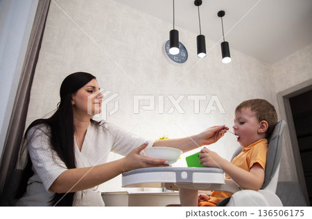Woman in bathrobe feeds young boy sitting in modern high chair inside bright home kitchen. Family mealtime activity with mother and child sharing healthy food. 136506175