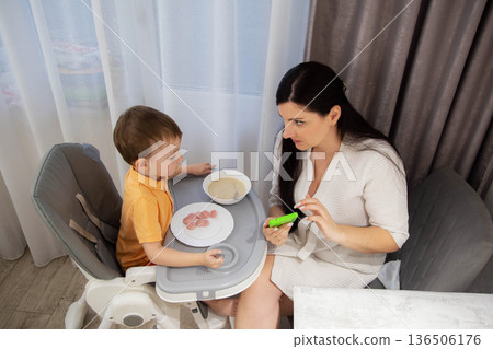 Young woman and small boy during mealtime indoors. Woman interacting with boy in high chair, boy has sausages and porridge on tray. Parenting scene at home. 136506176