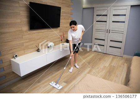 Woman mops wooden floor of modern home interior, performing routine household cleaning task with flat mop in spacious living area. 136506177