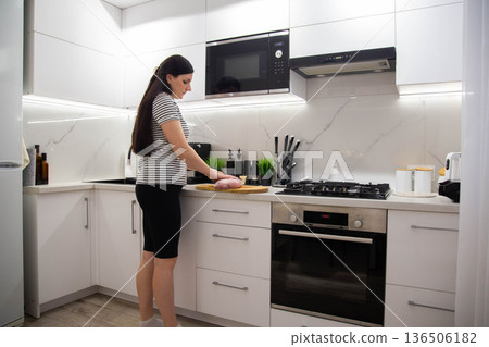 Woman stands in modern white kitchen, preparing raw meat on cutting board, day light illumination of contemporary home interior. Woman stands in modern white kitchen, preparing raw meat on cutting board, day light illumination of contemporary home interior. 136506182