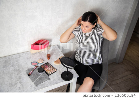 Young adult female sits at vanity table adjusting hair, surrounded by makeup products and mirror, preparing for daily routine or special occasion. Copy space for text Young adult female sits at vanity table adjusting hair, surrounded by makeup products and mirror, preparing for daily routine or special occasion. Copy space for text 136506184