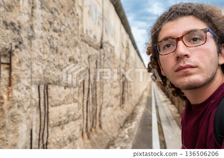 Berlin, germany, august 13, 2023. Young man in glasses gazing at the berlin wall memorial, reflecting on division, history and freedom beneath a cloudy sky Berlin, germany, august 13, 2023. Young man in glasses gazing at the berlin wall memorial, reflecting on division, history and freedom beneath a cloudy sky 136506206