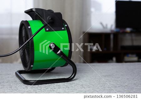 Electric heater with fan blows toward computer desk to warm person workspace at home. The challenges of working from home in cold weather. Raising room temperature with heat gun Electric heater with fan blows toward computer desk to warm person workspace at home. The challenges of working from home in cold weather. Raising room temperature with heat gun 136506281