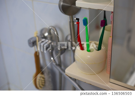 Close-up shelf with toothbrushes on bathroom wall near mirror above sink with faucet in background. Natural bathroom concept with worn look. Imperfect interior with toothbrushes in holder for family Close-up shelf with toothbrushes on bathroom wall near mirror above sink with faucet in background. Natural bathroom concept with worn look. Imperfect interior with toothbrushes in holder for family 136506503