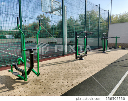 Outdoor exercise equipment in public park next to basketball court, modern fitness area with green and black machines, clear blue sky over sports facility, paved ground surface, daytime scene 136506530